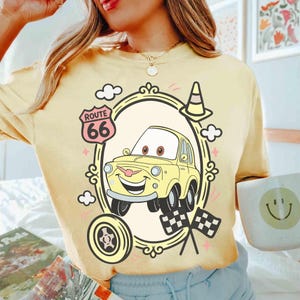 May include: Pale yellow t-shirt with a cartoon car design, Route 66 sign, traffic cone, and checkered flags. The car is within an oval frame with decorative elements. A wheel and clouds are also included in the design.