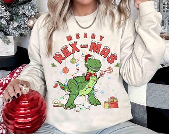 Puede incluir: Sudadera color crema con un gráfico festivo "Merry Rex-Mas". El diseño presenta un dinosaurio verde con gorro y bufanda de Papá Noel, adornado con luces navideñas y adornos. Una bolsa de regalo roja y una bola de nieve completan el tema navideño.