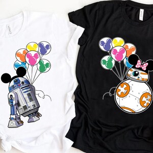 Puede incluir: Dos camisetas, una blanca y otra negra, cada una con un personaje de dibujos animados con orejas de Mickey Mouse sosteniendo globos de colores. La camiseta blanca tiene a R2-D2, y la camiseta negra a BB-8.