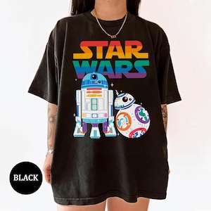 Puede incluir: Camiseta negra con el texto "STAR WARS" en colores arcoíris sobre ilustraciones de R2-D2 y BB-8. La camiseta tiene cuello redondo y mangas cortas. La palabra "BLACK" está en un círculo negro en la parte inferior.