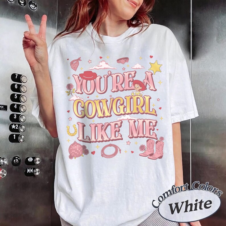 May include: A white short-sleeve t-shirt featuring the text "YOU'RE A COWGIRL LIKE ME" in pink and yellow. The graphic includes illustrations of a cowgirl hat, a cartoon character, a horseshoe, a disco ball, a lasso, and pink cowboy boots.