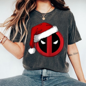 May include: Dark gray t-shirt featuring a red and black graphic of Deadpool's mask wearing a Santa hat. The design includes white eyes and a white trim on the hat. The shirt is worn by a person wearing blue jeans.