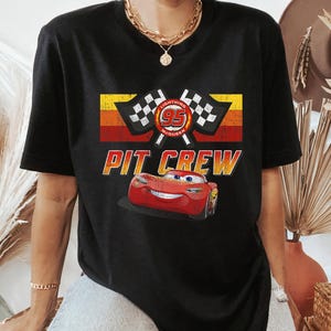 Peut inclure: T-shirt noir avec un motif de Flash McQueen du film Cars. Le design comprend le numéro 95, des drapeaux à damier et les mots "PIT CREW" dans un style rétro. Le t-shirt a un col rond classique.