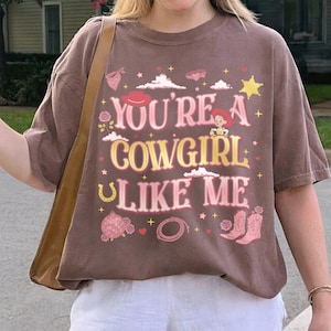 May include: A brown short-sleeve t-shirt featuring the text "YOU'RE A COWGIRL LIKE ME" in pink and yellow. The graphic includes illustrations of a cowgirl, a cowboy hat, a star, a horseshoe, a disco ball, a lasso, and pink cowboy boots.