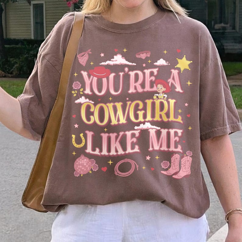 May include: A brown short-sleeve t-shirt featuring the text "YOU'RE A COWGIRL LIKE ME" in pink and yellow. The graphic includes illustrations of a cowgirl, a cowboy hat, a star, a horseshoe, a disco ball, a lasso, and pink cowboy boots.