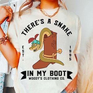 Op de afbeelding: Crèmekleurig T-shirt met een cartoon slang met hoed, die uit een bruine cowboylaars komt. De tekst luidt "THERE'S A SNAKE IN MY BOOT" met "EST. 1995" en "WOODY'S CLOTHING CO."