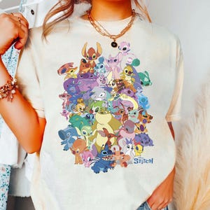 Könnte beinhalten: Cremefarbenes T-Shirt mit einer farbenfrohen Grafik verschiedener Stitch-Charaktere aus dem Disney-Film. Das Design zeigt Stitch in verschiedenen Posen und Farben, mit dem Wort "Stitch" in einer stilisierten Schriftart unten. Das Shirt hat kurze Ärmel.