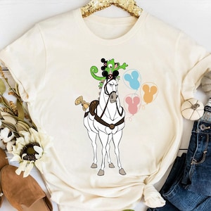 May include: Cream-colored t-shirt featuring a cartoon illustration of a white horse with a frog wearing a Mickey Mouse hat on its back. The horse is holding three Mickey Mouse-shaped balloons in blue, pink, and orange.