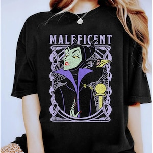 Puede incluir: Camiseta negra con un diseño de Maléfica, un cuervo y un bastón, enmarcado por elementos decorativos y la palabra "Maléfica". El diseño utiliza verde, morado, amarillo y blanco.