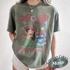 May include: Olive green t-shirt featuring the words "Aloha From" above a cartoon illustration of Lilo and Stitch in a tropical setting. The shirt also has the text "Kaua'i Town, HI" and the words "Comfort Colors Moss" in an oval.