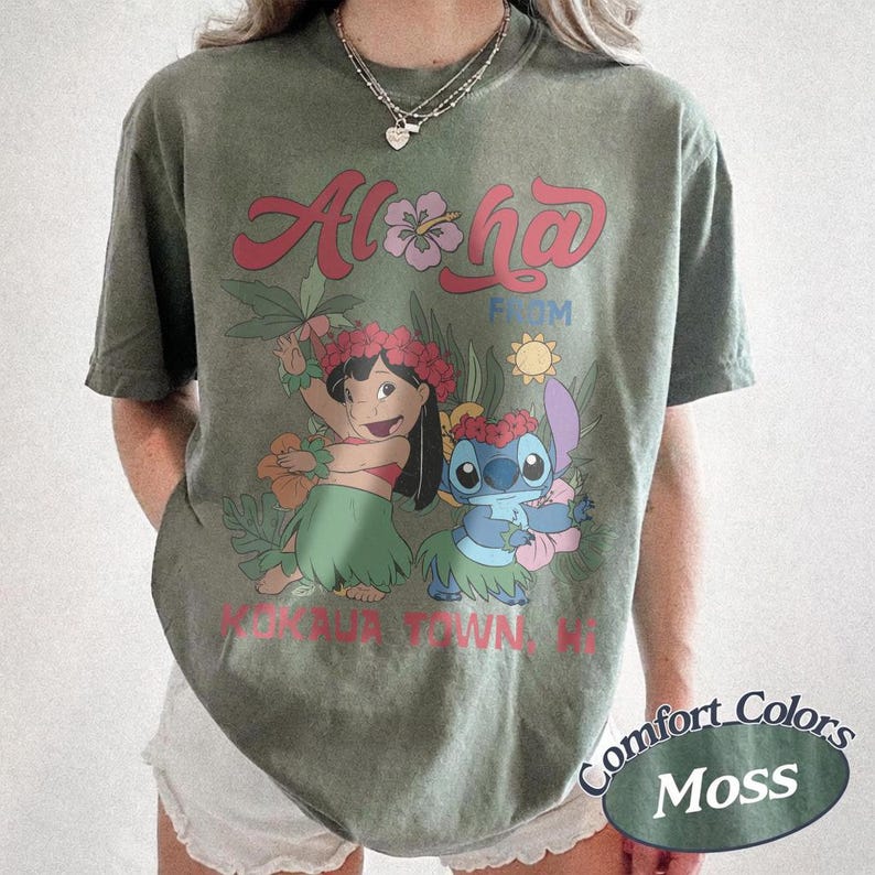 May include: Olive green t-shirt featuring the words "Aloha From" above a cartoon illustration of Lilo and Stitch in a tropical setting. The shirt also has the text "Kaua'i Town, HI" and the words "Comfort Colors Moss" in an oval.