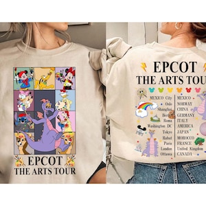 Puede incluir: Sudadera beige con un gráfico colorido de personajes de Disney y el texto "EPCOT THE ARTS TOUR". La parte trasera de la sudadera muestra el texto "EPCOT THE ARTS TOUR" con una lista de ciudades y una ilustración de un dragón morado.