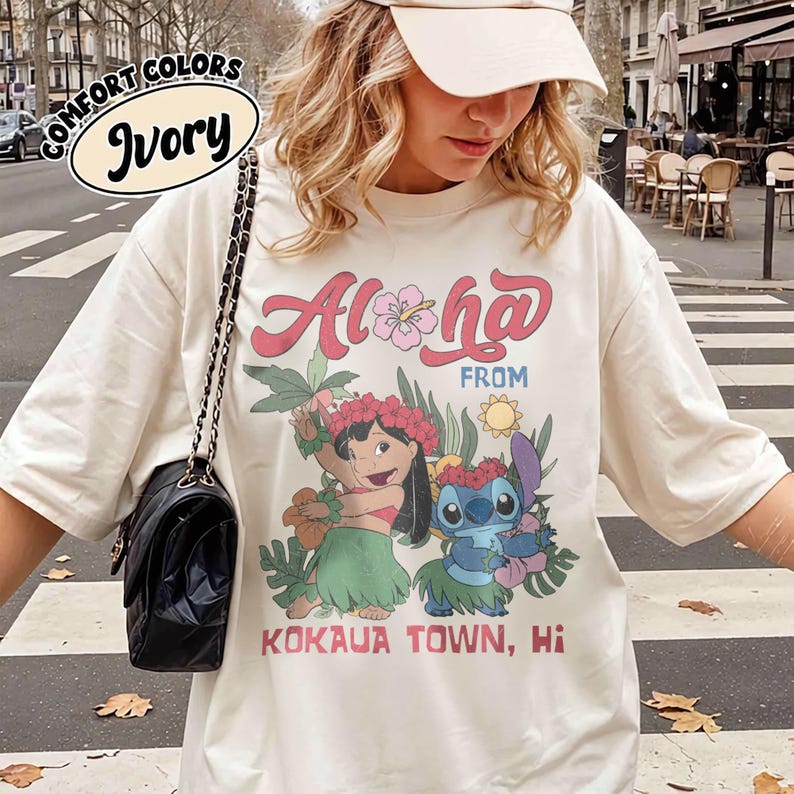 May include: Ivory-colored t-shirt with the words "Aloha From Kokaua Town, Hi" in pink and red lettering. The shirt features a graphic of two cartoon characters with floral accents and tropical foliage.