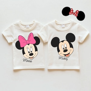 以下が含まれることがあります： ミッキーとミニーマウスが描かれた2枚の白いTシャツ。ミニーのシャツにはピンクのリボンと画像の下に「Minnie」という名前があります。ミッキーのシャツには画像の下に「Mickey」という名前があります。赤い水玉模様のリボンが付いたミッキーマウスの耳のペアも含まれています。