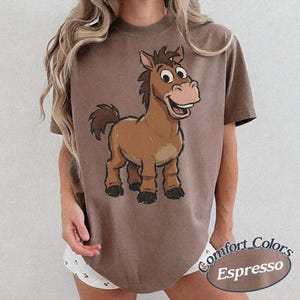 May include: A brown short-sleeve t-shirt featuring a cartoon illustration of a brown horse with a dark mane and tail. The horse has large eyes and a wide smile. The text Comfort Colors Espresso is printed in a dark oval graphic at the bottom right.