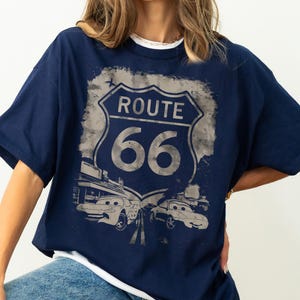May include: A navy blue short-sleeve t-shirt featuring a distressed graphic of a Route 66 road sign. Below the sign, two cartoon-style cars with expressive eyes are depicted on a road, set against a faded, textured background design.