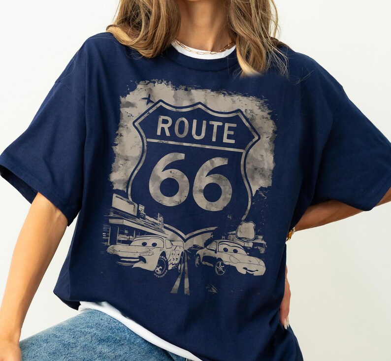 May include: A navy blue short-sleeve t-shirt featuring a distressed graphic of a Route 66 road sign. Below the sign, two cartoon-style cars with expressive eyes are depicted on a road, set against a faded, textured background design.