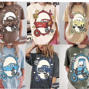 May include: Six t-shirts in different colours, each with a cartoon car design inside a Route 66 themed frame. The shirts are in shades of brown, cream, yellow, blue, grey, and green. The text "Route 66" is visible on each shirt.