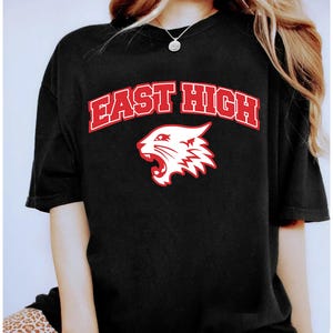 May include: Black t-shirt with the text "EAST HIGH" in red block letters with a white outline. Below the text is a white and red graphic of a wildcat's head with its mouth open.