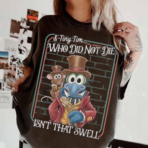 Puede incluir: Camiseta gris oscuro con un gráfico de Gonzo el Grande y un ratón, con el texto "& Tiny Tim... WHO DID NOT DIE ISN'T THAT SWELL" en un marco rectangular. Gonzo lleva un sombrero de copa y un abrigo rojo.