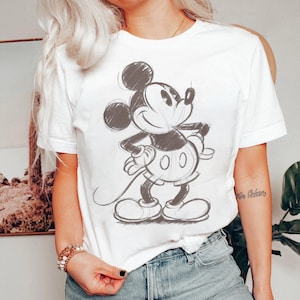 Könnte beinhalten: Weißes T-Shirt mit einer schwarz-grauen Skizze von Mickey Mouse. Die Cartoonfigur lächelt und hat die Hände in die Hüften gestemmt. Das Shirt ist ein klassischer Rundhalsausschnitt.