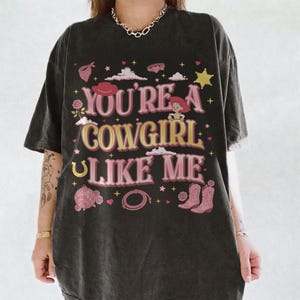 May include: A charcoal gray short-sleeve t-shirt featuring the text "YOU'RE A COWGIRL LIKE ME" in pink and yellow. The graphic includes illustrations of a cowboy hat, a cartoon character, a disco ball, a lasso, boots, stars, and clouds.