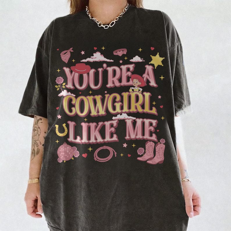 May include: A charcoal gray short-sleeve t-shirt featuring the text "YOU'RE A COWGIRL LIKE ME" in pink and yellow. The graphic includes illustrations of a cowboy hat, a cartoon character, a disco ball, a lasso, boots, stars, and clouds.