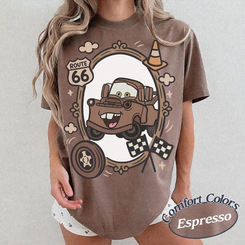 May include: Espresso-coloured t-shirt featuring a cartoon image of Mater from the movie Cars, framed by a decorative oval. The design includes Route 66 signage, a traffic cone, and checkered flags. The shirt has a Comfort Colors label.