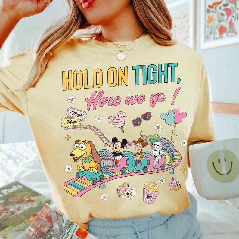 Op de afbeelding: Lichtgeel T-shirt met de tekst "HOLD ON TIGHT, Here we go!" en een cartoon trein met Slinky Dog, Mickey Mouse, Woody en een Stormtrooper. Het ontwerp bevat ijs, zonnebrillen en popcorn.