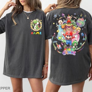 Pu&ograve; includere: T-shirt grigio scuro con una grafica colorata dei personaggi del gioco Super Mario Galaxy sul retro. La parte anteriore presenta un design del personaggio Yoshi con la scritta "NAME" in colori arcobaleno. La maglietta ha una vestibilit&agrave; ampia.