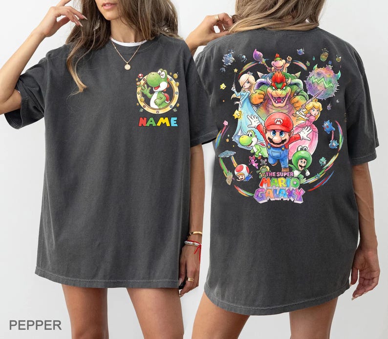 Pu&ograve; includere: T-shirt grigio scuro con una grafica colorata dei personaggi del gioco Super Mario Galaxy sul retro. La parte anteriore presenta un design del personaggio Yoshi con la scritta "NAME" in colori arcobaleno. La maglietta ha una vestibilit&agrave; ampia.