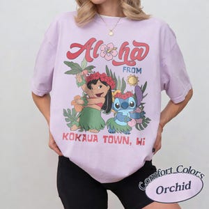 May include: Orchid-colored t-shirt featuring a graphic of Lilo and Stitch with the text "Aloha From Kokaua Town, Hi." The design includes floral elements and the Comfort Colors logo.