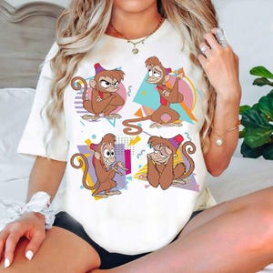 Op de afbeelding: Crèmekleurig T-shirt met een cartoonontwerp van vier apen met verschillende uitdrukkingen. De apen zijn bruin met rode en paarse hoeden. De achtergrond bevat kleurrijke geometrische vormen in roze, blauw en geel.