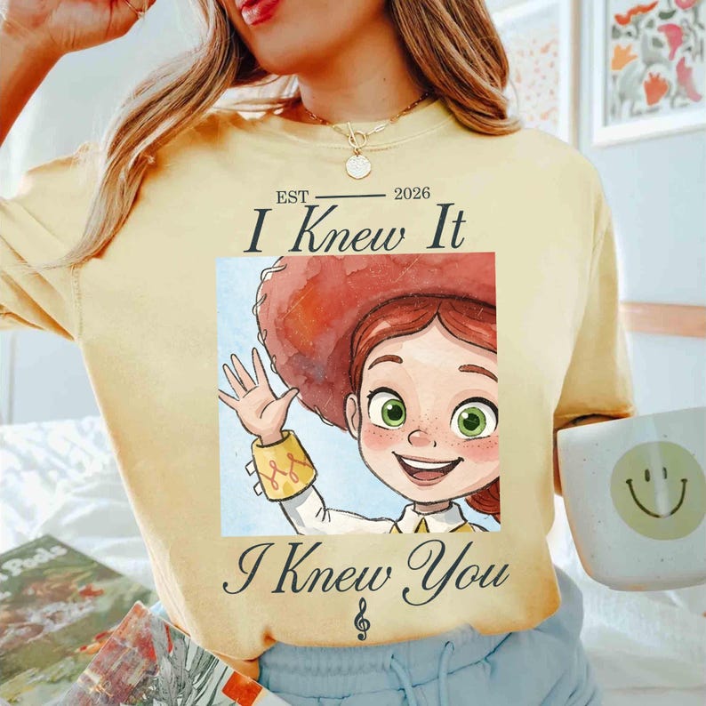 Peut inclure: Un t-shirt jaune p&acirc;le avec une illustration de personnage de dessin anim&eacute; aux cheveux roux portant un chapeau de cow-boy et faisant un signe de la main. Le texte indique "EST 2026 I Knew It I Knew You" avec une note de musique en dessous.