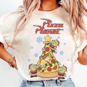 May include: Cream-colored t-shirt featuring a festive Pizza Planet design. The graphic includes a pizza slice decorated as a Christmas tree, with three green aliens in Santa suits. The words "Pizza Planet" are written in red with a snowy effect.
