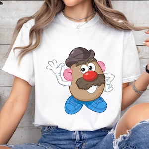 May include: White t-shirt with a cartoon Mr. Potato Head design. The character has a brown hat, mustache, and a red nose. The design includes blue shoes and pink ears. This casual t-shirt is suitable for everyday wear.