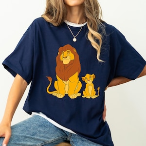 May include: Navy blue t-shirt featuring a cartoon illustration of a lion and a lion cub. The lion has a brown mane and the cub is yellow. The shirt has a white collar and is worn with blue jeans.