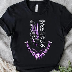 Könnte beinhalten: Schwarzes T-Shirt mit einer weißen und lila Grafik der Black Panther-Maske. Die Worte "WAKANDA FOREVER" sind vertikal links neben der Maske ausgerichtet. Ein lila halskettenartiges Design befindet sich unten.
