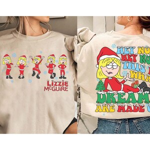 Pode incluir: Camisola bege com um desenho de desenho animado de Lizzie McGuire com um chapéu de Papai Noel e uma roupa vermelha. A frente mostra Lizzie em várias poses, e as costas têm o texto "Hey Now, This Is What Dreams Are Made Of."