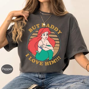 May include: Dark gray t-shirt featuring a distressed graphic of Ariel from The Little Mermaid. The design includes Ariel with red hair, a blue top, and green tail, surrounded by text that reads "BUT DADDY I LOVE HIM!!!".