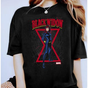 May include: Black t-shirt featuring a comic book style illustration of Black Widow. The design includes a red triangle, a spiderweb background, and the text "BLACK WIDOW" in red and yellow. The Marvel logo is at the bottom.