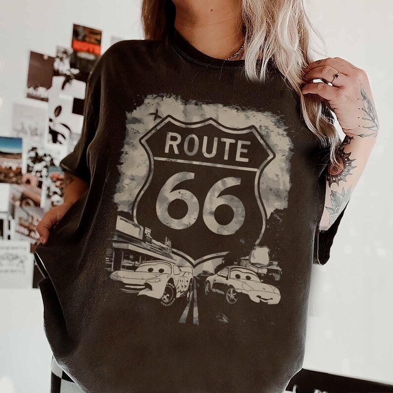 May include: A charcoal gray short-sleeve t-shirt featuring a distressed graphic of the Route 66 highway shield sign. Below the sign, the design includes two cartoon-style race cars on a road, with the text ROUTE 66 printed in bold white letters.