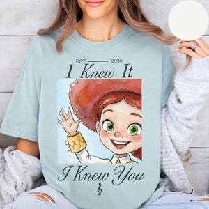 Peut inclure: Un t-shirt bleu clair avec une illustration d'un personnage de dessin anim&eacute; aux cheveux roux portant un chapeau rouge, faisant un signe de la main. Le texte indique EST 2026, I Knew It, I Knew You, avec une note de musique en dessous.