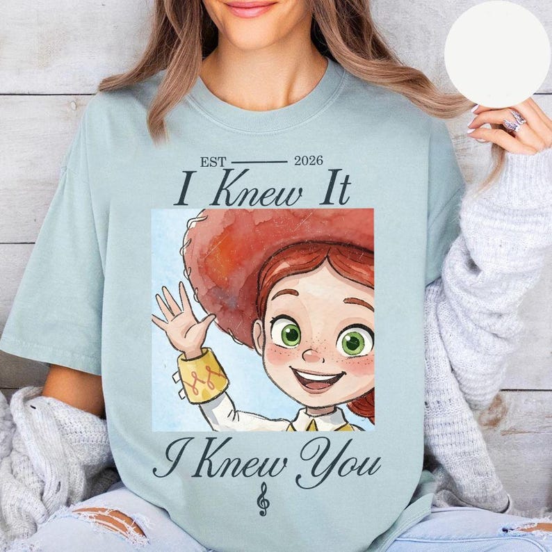 Peut inclure: Un t-shirt bleu clair avec une illustration d'un personnage de dessin anim&eacute; aux cheveux roux portant un chapeau rouge, faisant un signe de la main. Le texte indique EST 2026, I Knew It, I Knew You, avec une note de musique en dessous.
