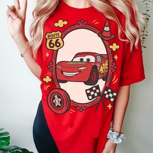 May include: Red t-shirt with a cartoon Lightning McQueen design. The design includes a Route 66 sign, a racing cone, and checkered flags. The shirt is a bright red colour.
