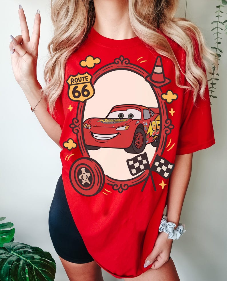 May include: Red t-shirt with a cartoon Lightning McQueen design. The design includes a Route 66 sign, a racing cone, and checkered flags. The shirt is a bright red colour.