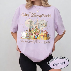 K&ouml;nnte beinhalten: Helllila T-Shirt mit einem Aufdruck mit den Worten "Walt Disney World" und "The Happiest Place on Earth". Die Grafik zeigt verschiedene Disney-Figuren und ein Schloss. Das Shirt ist mit "Comfort Colors Orchid" gekennzeichnet.