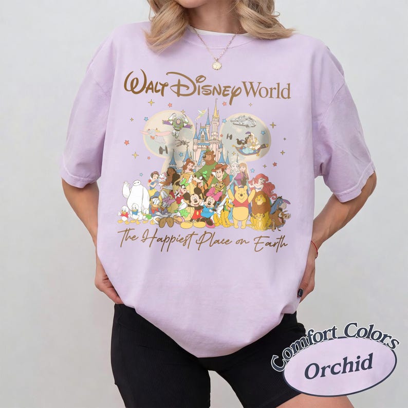 K&ouml;nnte beinhalten: Helllila T-Shirt mit einem Aufdruck mit den Worten "Walt Disney World" und "The Happiest Place on Earth". Die Grafik zeigt verschiedene Disney-Figuren und ein Schloss. Das Shirt ist mit "Comfort Colors Orchid" gekennzeichnet.