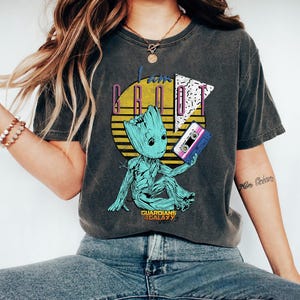 May include: Dark gray t-shirt featuring a teal Groot character holding a cassette tape. The shirt has the text "I am Groot" in a retro design with a yellow sunburst and the words "Guardians of the Galaxy Vol. 2".