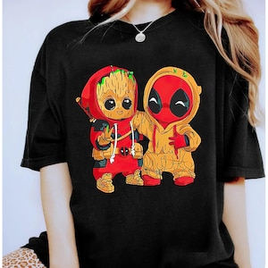 May include: Black t-shirt featuring a cartoon illustration of two characters in hoodies. One is red with a Deadpool mask, the other is brown with a Groot-like design. The characters are embracing, with one giving a peace sign.
